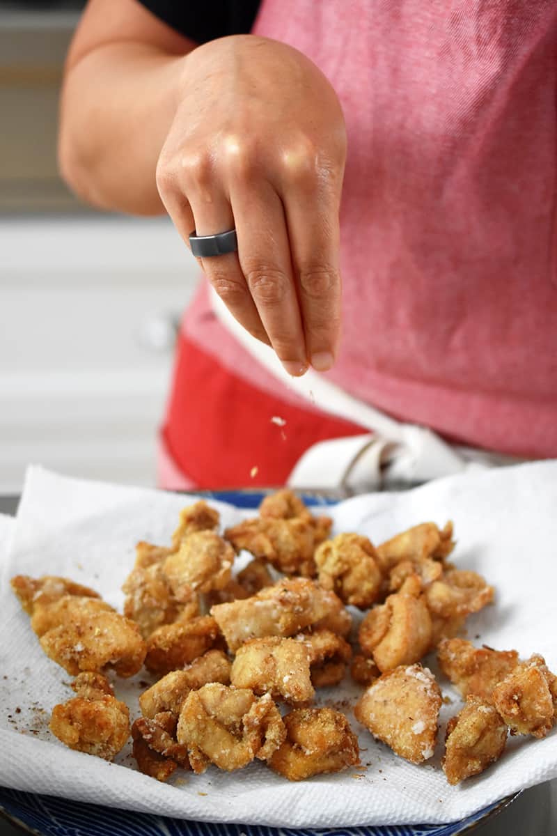 A person in a red apron is sprinkling crushed Sichuan peppercorns and salt on gluten-free Taiwanese popcorn chicken.