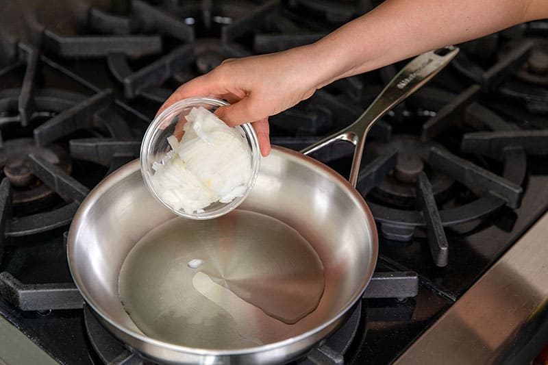 Adding diced onions to a stainless steel skillet with olive oil to make the sauce for eggs in purgatory.