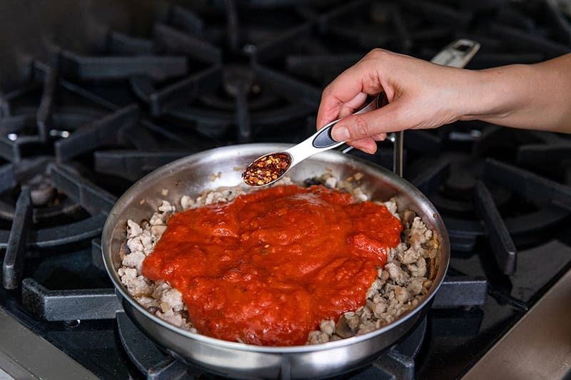 A hand adding spoonful of crushed red peppers to a stainless steel skillet filled with cooked sausage, mushrooms, and marinara sauce.