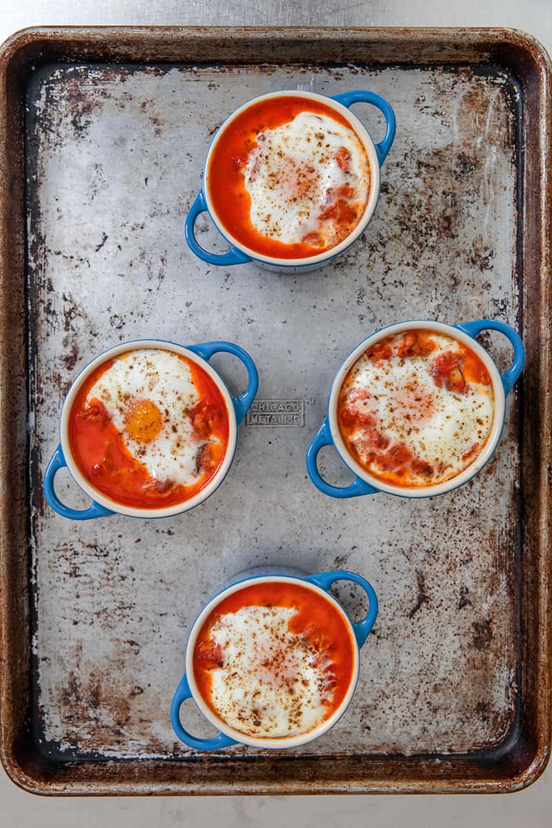 An overhead shot of a rimmed baking sheet topped with four blue cocottes filled with paleo eggs in purgatory right out of the oven. 