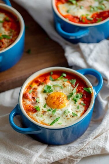An overhead shot of three blue cocottes filled with Whole30 eggs in purgatory, baked eggs in a spicy tomato meat sauce