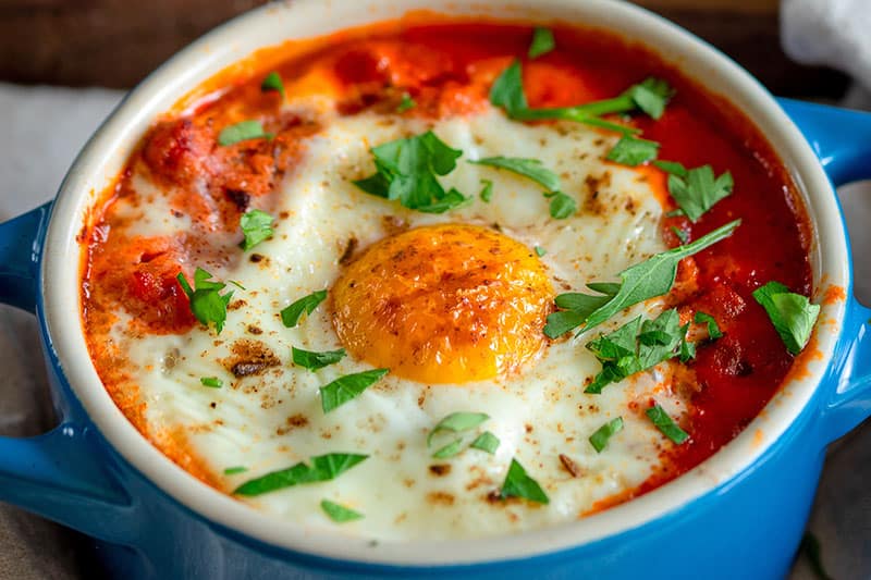 A closeup shot of a blue cocotte filled with eggs in purgatory topped with minced Italian parsley.