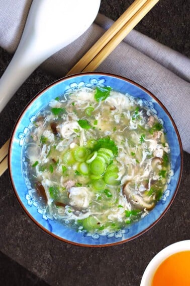 An overhead shot of West Lake Beef Soup in a blue bowl next to a pair of chopsticks and a white ceramic soup spoon.