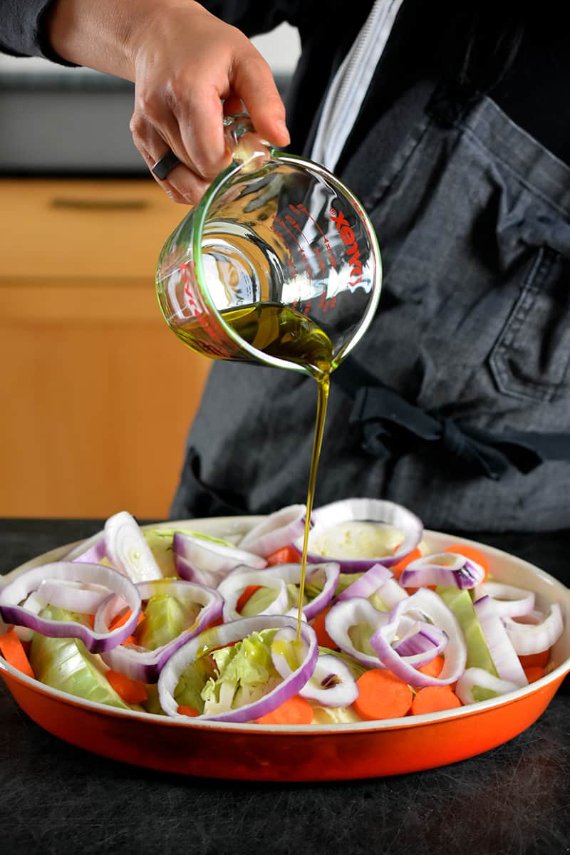 Someone in a gray apron is pouring olive oil from a liquid measuring cup onto a casserole pan filled with cabbage wedges, carrots, and onions.