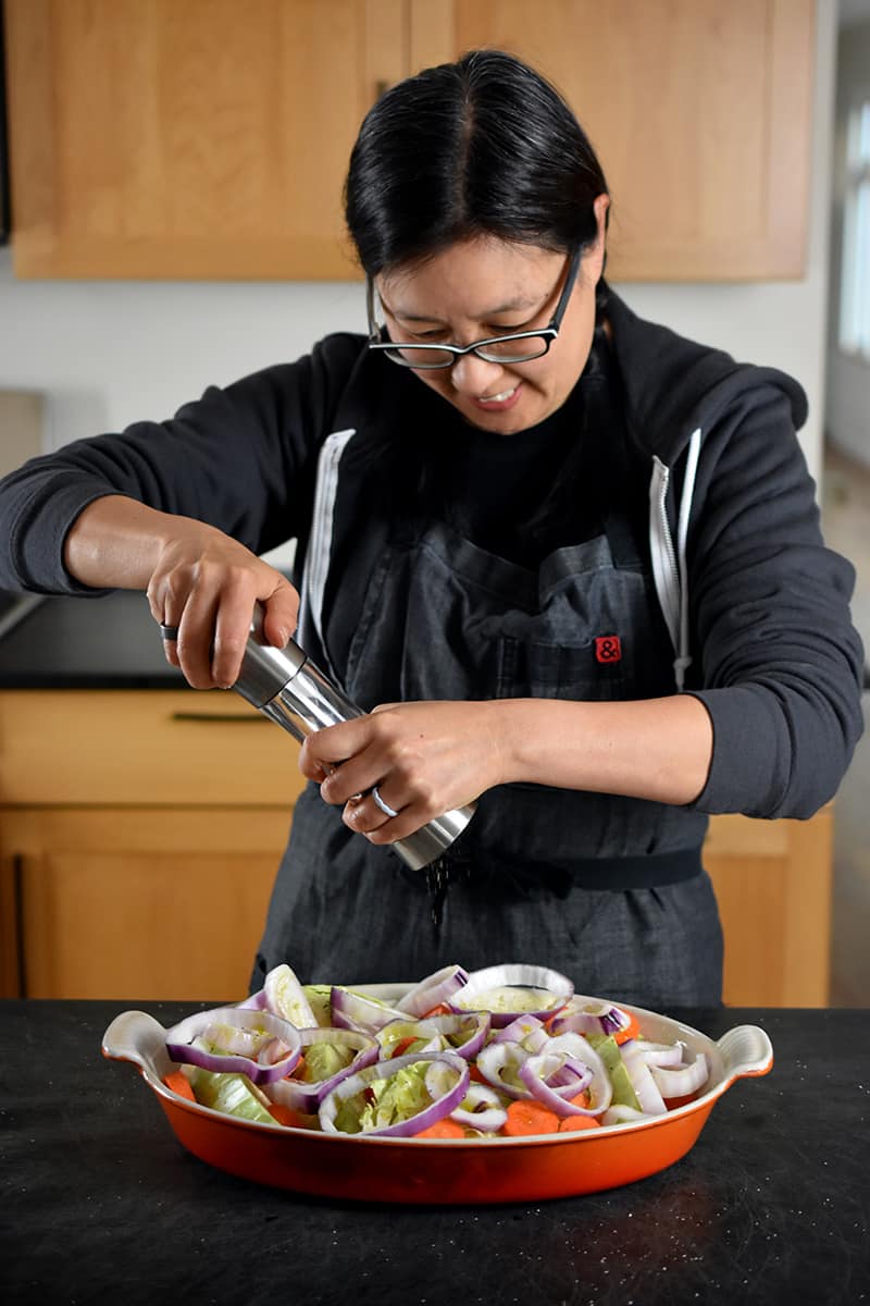 An Asian woman in glasses is adding freshly ground black pepper from a Peppermill to a casserole pan filled with cabbage wedges, carrots, and onions.