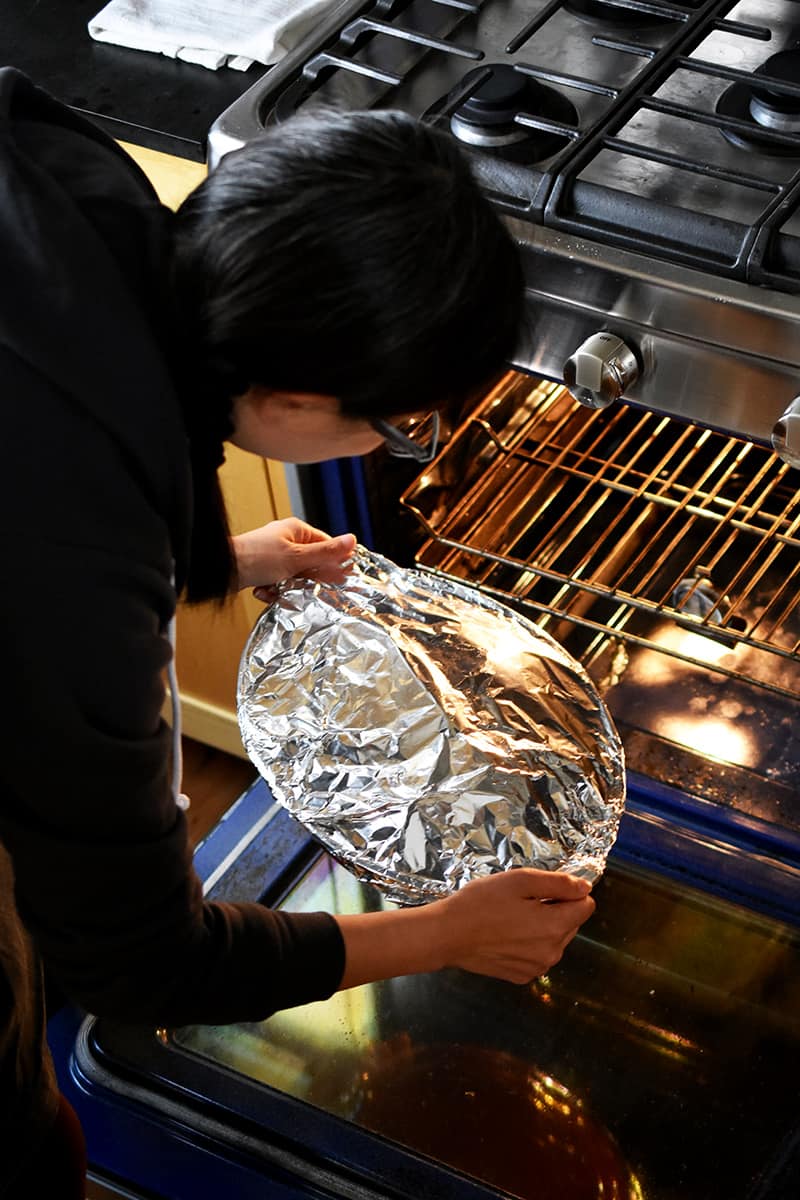 A brunette woman is placing a foil covered casserole pan filled with braised cabbage into an open oven