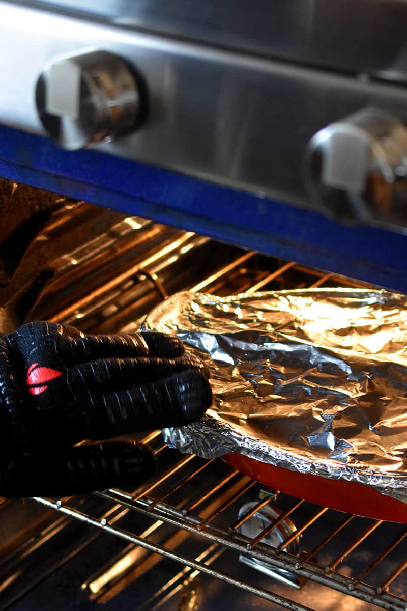 A gloved hand is placing a foil covered casserole filled with braised cabbage in an open oven