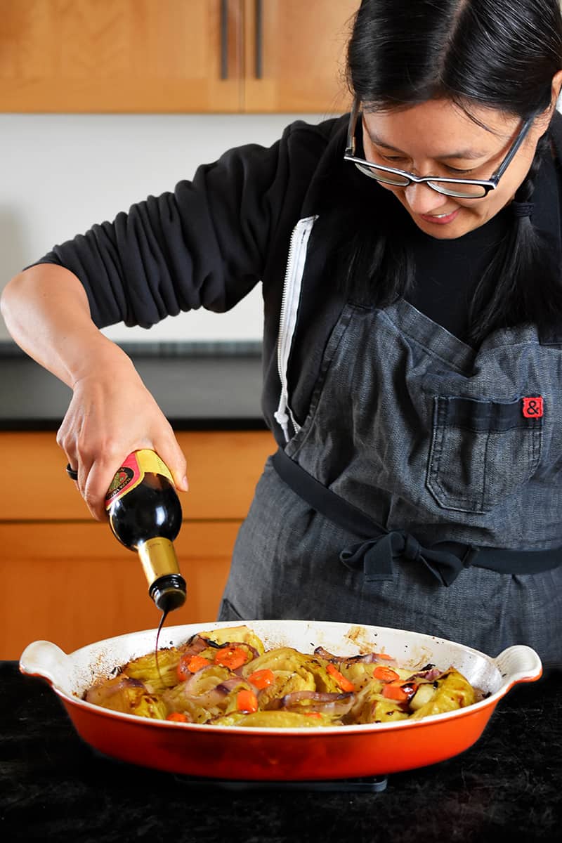 An Asian woman in a gray apron is pouring balsamic vinegar on braised cabbage right out of the oven.