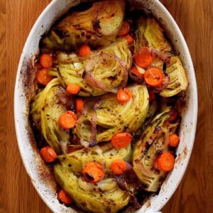 An overhead shot of the world's best braised green cabbage, carrot coins, and sliced onions in an oval casserole pan. Two hands are holding the handles with towels.
