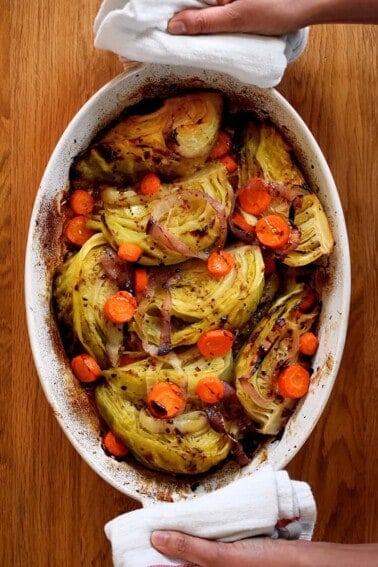 An overhead shot of the world's best braised green cabbage, carrot coins, and sliced onions in an oval casserole pan. Two hands are holding the handles with towels.