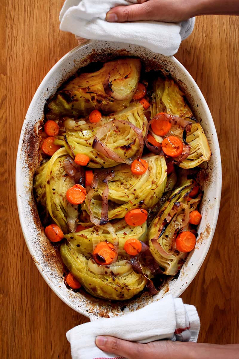 An overhead shot of the world's best braised green cabbage, carrot coins, and sliced onions in an oval casserole pan. Two hands are holding the handles with towels.