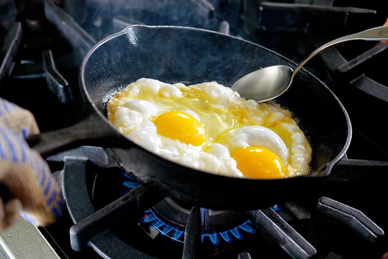 A cast iron skillet filled with two sunnyside up fried eggs is tilted with a spoon ladling hot ghee on the uncooked whites.