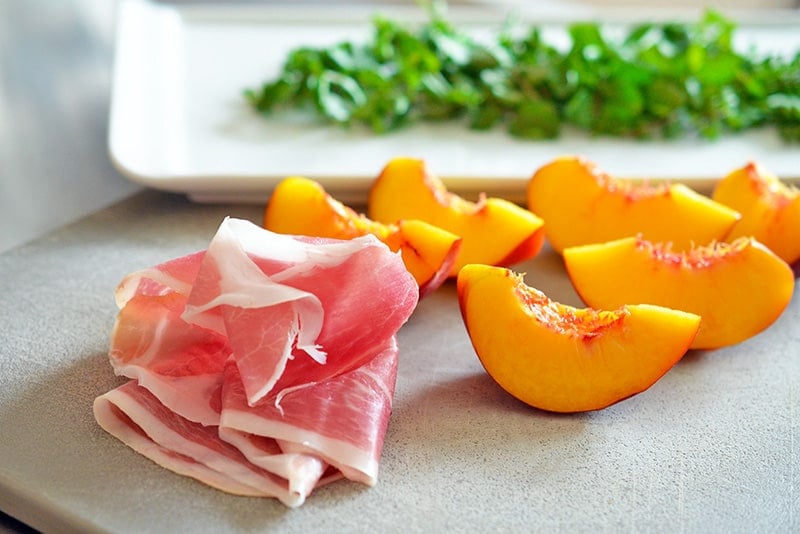 Quartered peaches, a slice of prosciutto, and washed watercress sitting on a cutting board.