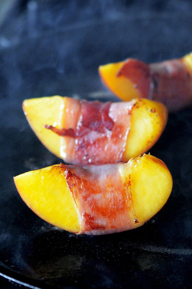 A close up of prosciutto-wrapped peached searing on a cast iron skillet.