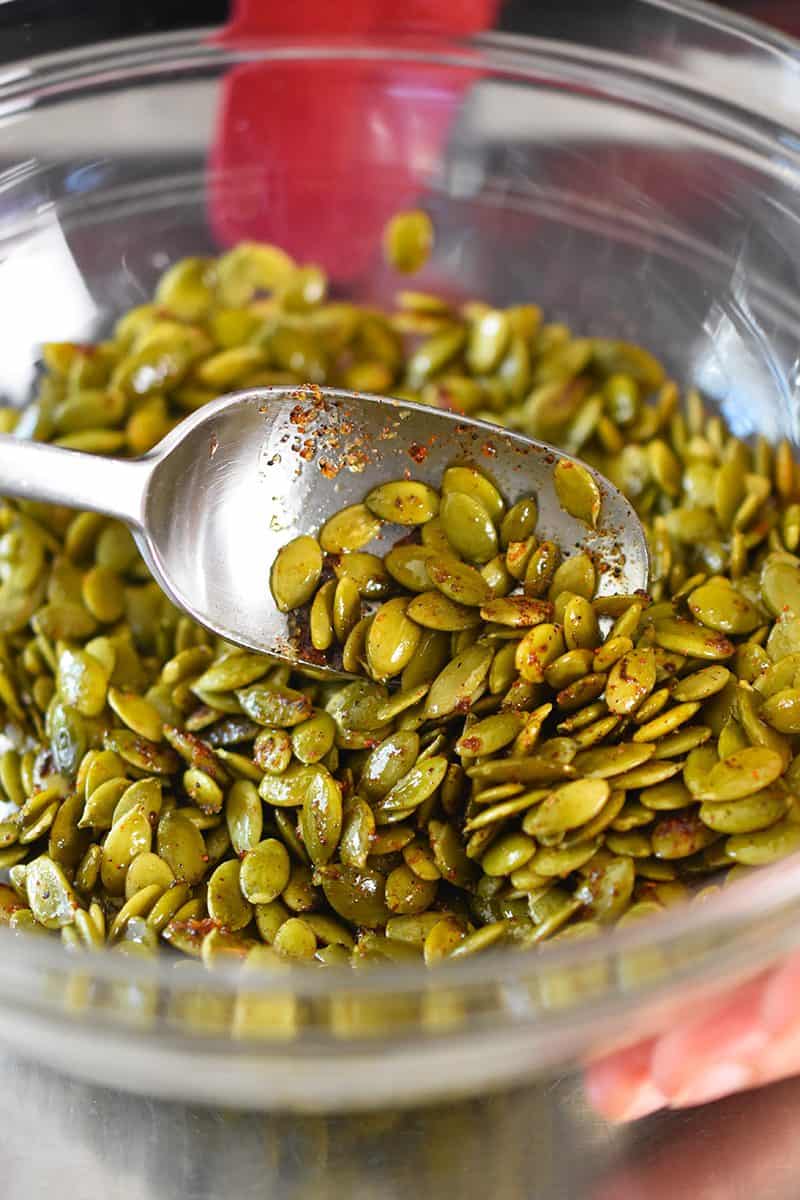A bowl of spiced pepitas with a spoon mixing the olive oil, salt, paprika, cayenne, and lime juice.