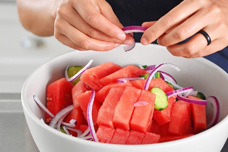 Someone breaking up the red onions to be put in a large white bowl with watermelons and cucumbers.