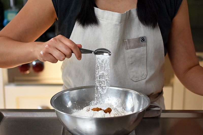 Someone adding a spoonful of baking soda to a bowl filled with the dry ingredients for apple Bundt Cake.