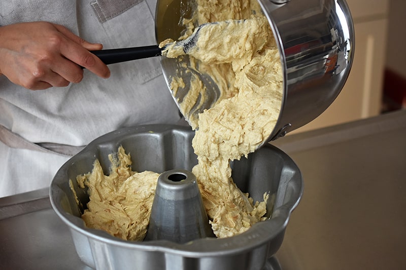 Someone using a black silicone spatula to transfer the apple Bundt Cake batter into a greased bundt pan.