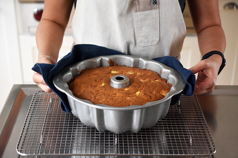 Someone using a towel to place a freshly baked apple Bundt Cake on a wire rack to cool in the pan.