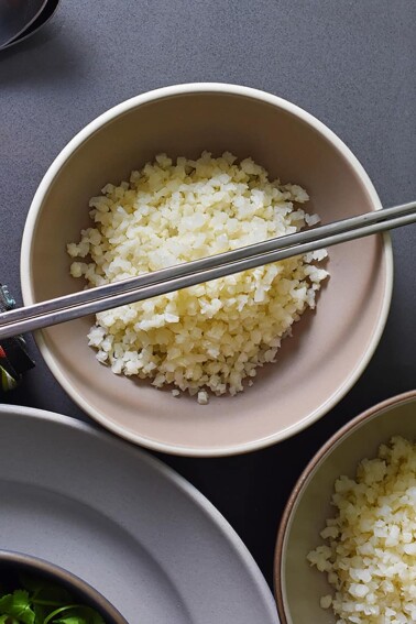 An overhead shot of cauliflower rice in a bowl topped with silver chopsticks