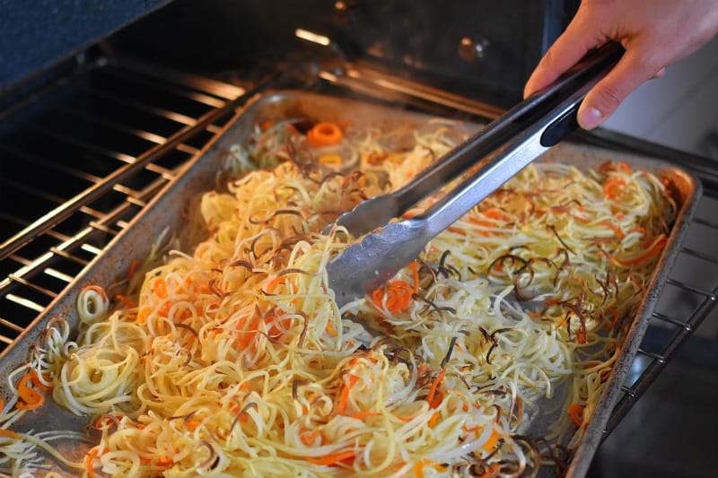 Crispy Swoodles being tossed by a pair of tongs as it cooks in the oven.