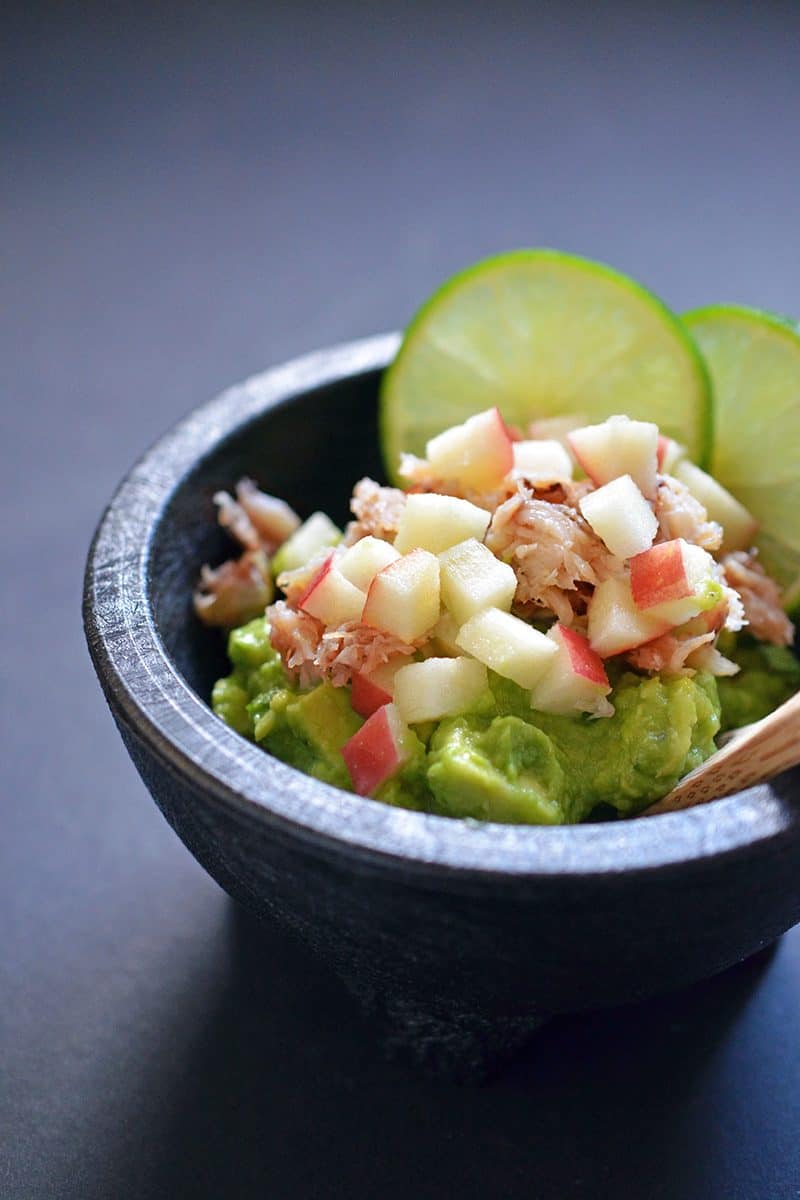 Guacamole in a black mocajete topped with lump crab meat, diced apples, and lime slices.