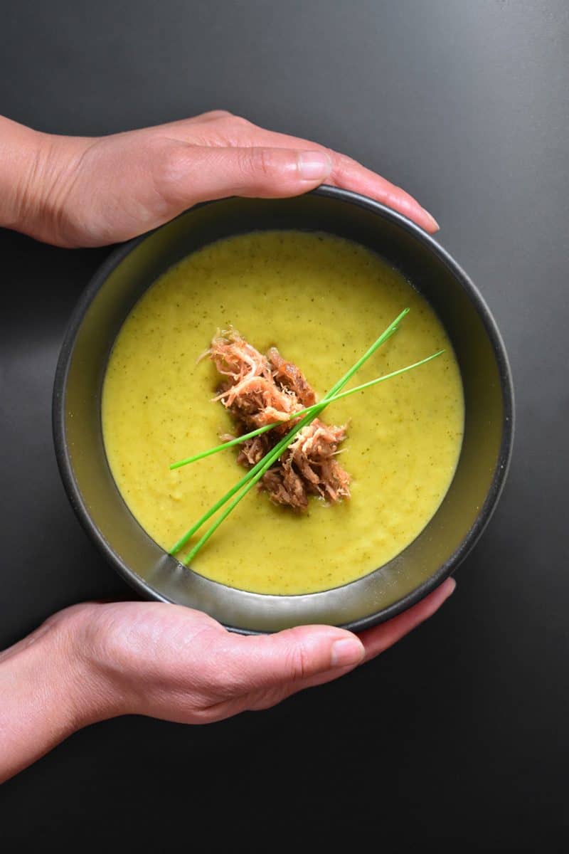 Someone holding a bowl of Instant Pot curried cream of broccoli soup topped with kalua pig.