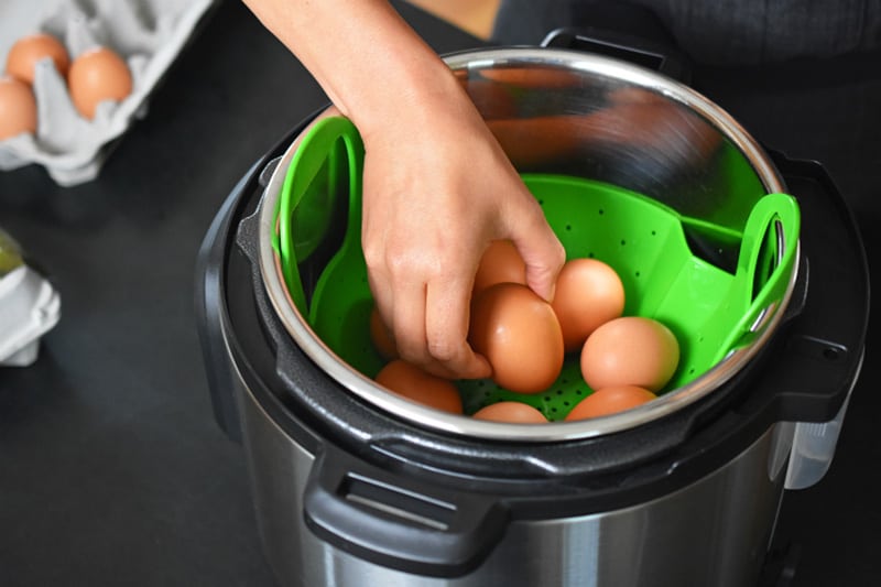A hand is shown placing eggs onto a steamer insert in an Instant Pot.