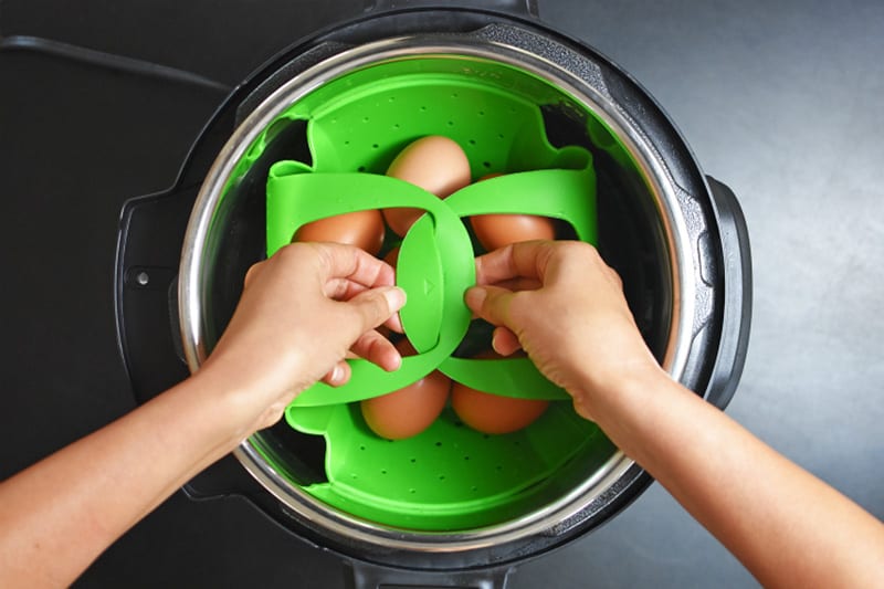 Two hands are shown locking the handles of the silicone steamer together in an Instant Pot.