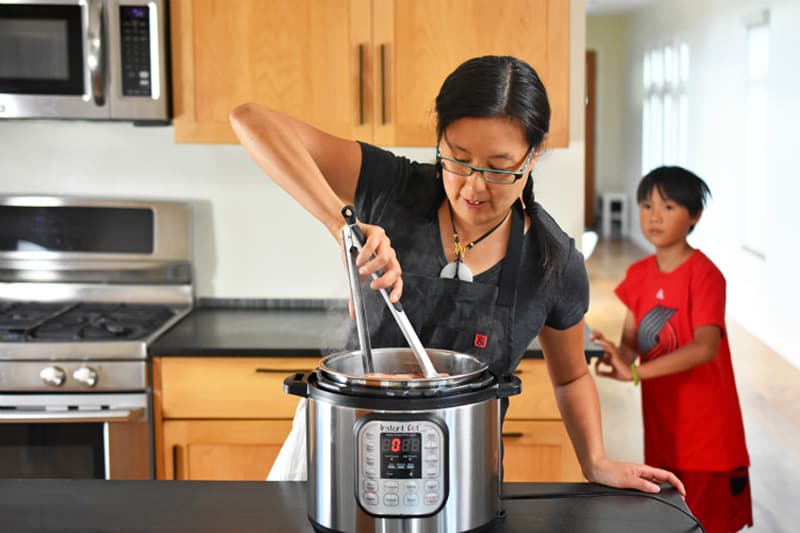 An Asian woman is using tongs to stir Instant Pot Summer Italian Chicken while a small boy watches.
