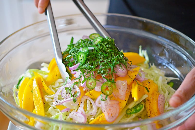 Someone tossing the spicy mango cabbage slaw with metal tongs in a large glass bowl.