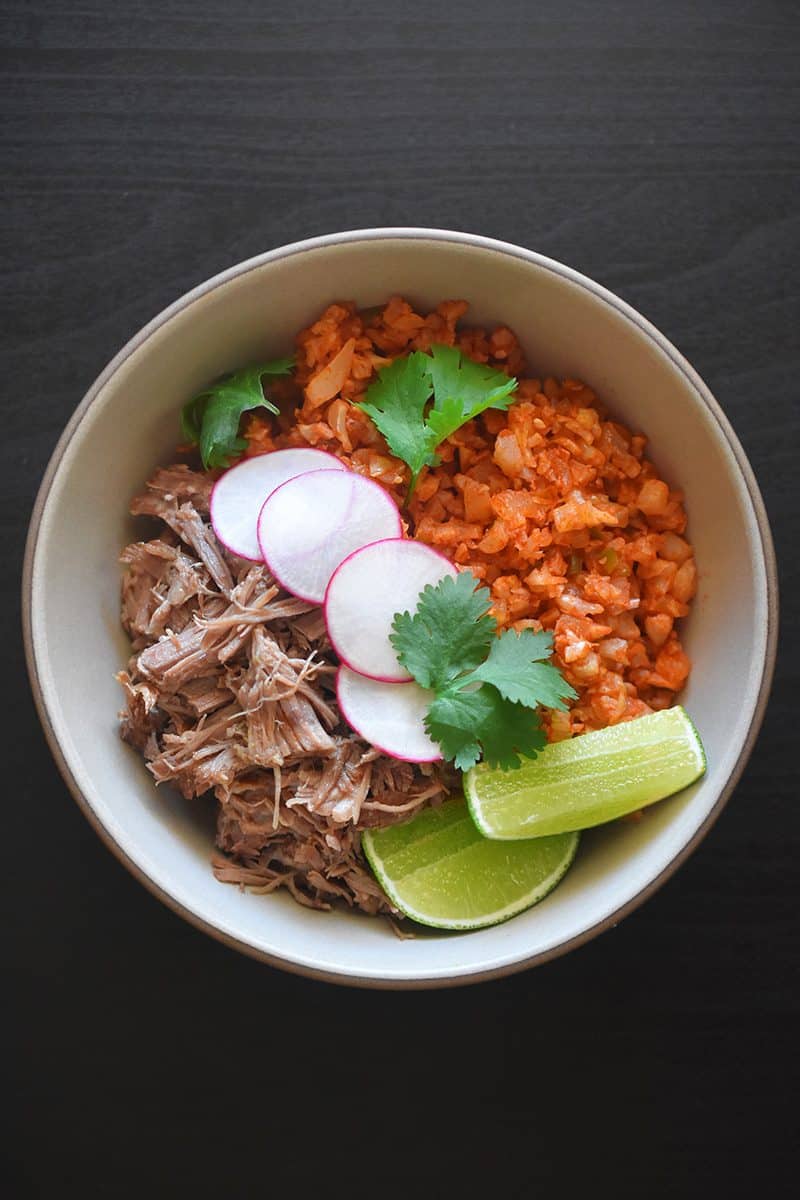An overhead shot of a Whole30 burrito bowl with Mexican Cauliflower Rice, carnitas, sliced limes, and radishes.