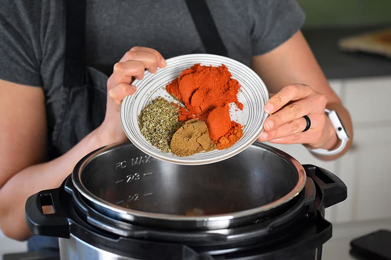 A closeup of a woman adding a plate filled with chili powder, oregano, cumin, and cayenne pepper into an open Instant Pot.