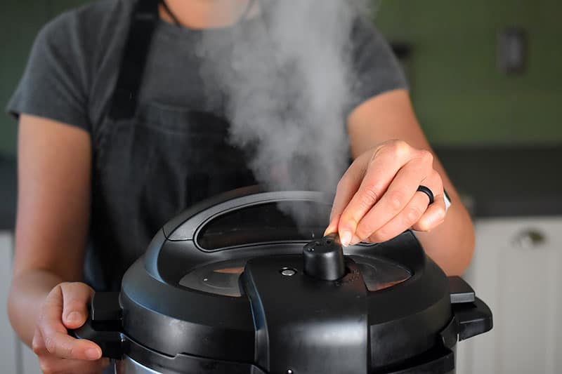 A closeup of a woman turning the pressure release valve on the lid of an Instant Pot. There is a stream of steam escaping from the valve.