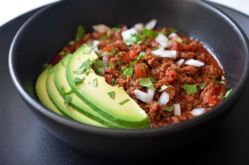 A side view of a black bowl on top of a black plate. The bowl is filled with Instant Pot Ground Beef Chili and topped with sliced avocado, diced white onions, and minced cilantro leaves.