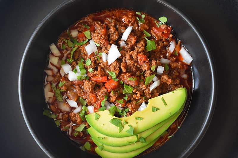 An overhead shot of a bowl of Instant Pot Ground Beef Chili topped with sliced avocado and diced white onions