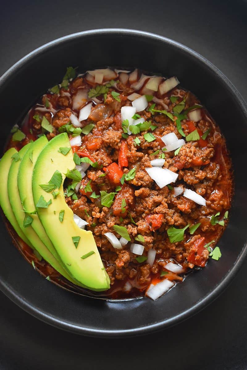 A top view of a black bowl on top of a black plate. The bowl is filled with Instant Pot Ground Beef Chili and topped with sliced avocado, diced white onions, and minced cilantro leaves.