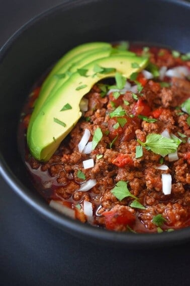 A closeup image of a bowl of Instant Pot Ground Beef Chili topped with sliced avocado, diced onions, and minced cilantro.