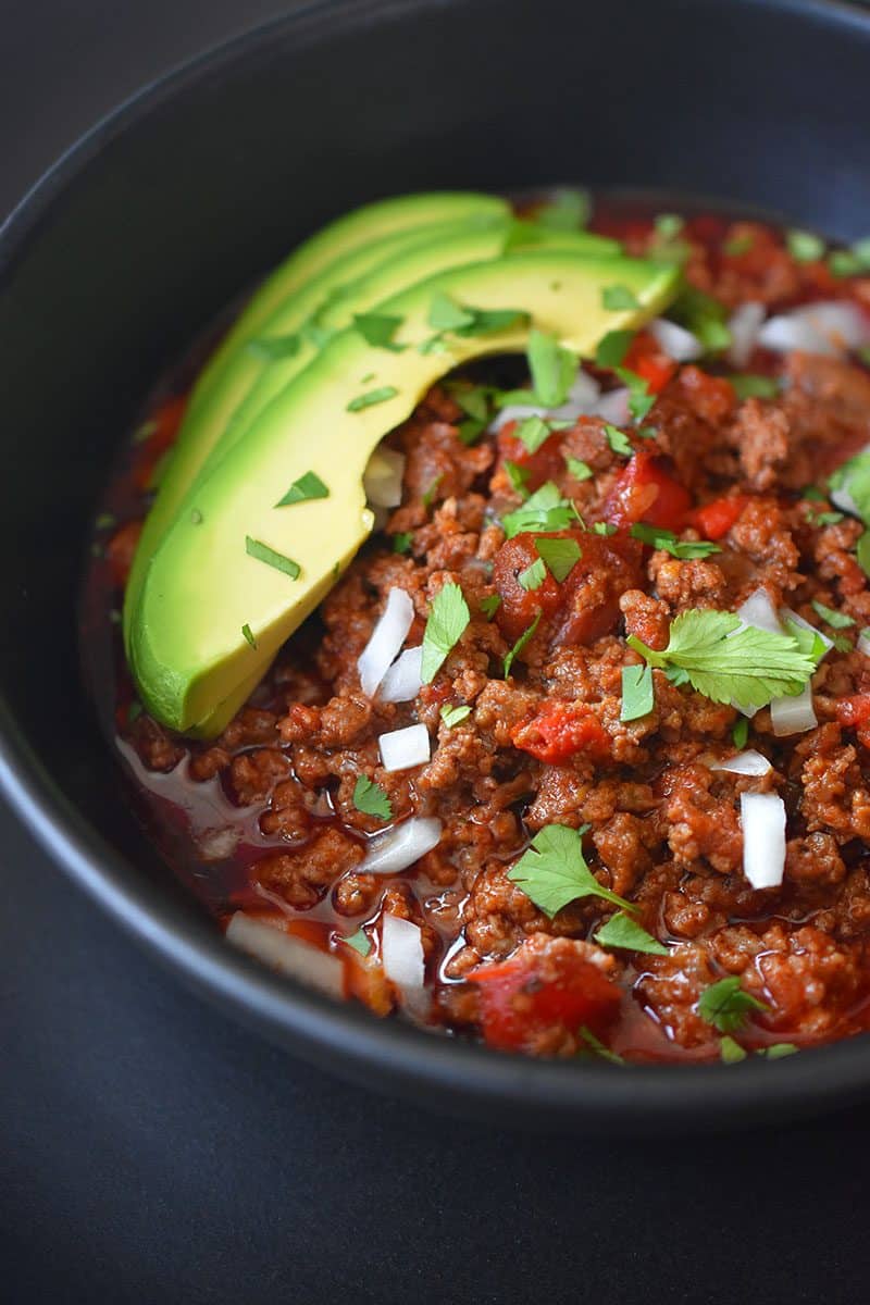 A close up image of Instant Pot Ground Beef Chili topped with sliced avocado, diced onions, and chopped cilantro in a black bowl.