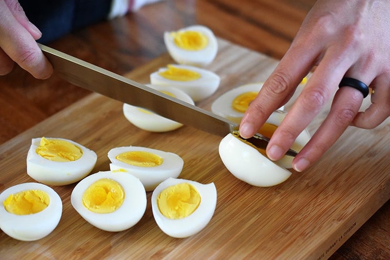 Someone slicing a bunch of hard cooked eggs on a wooden cutting board.