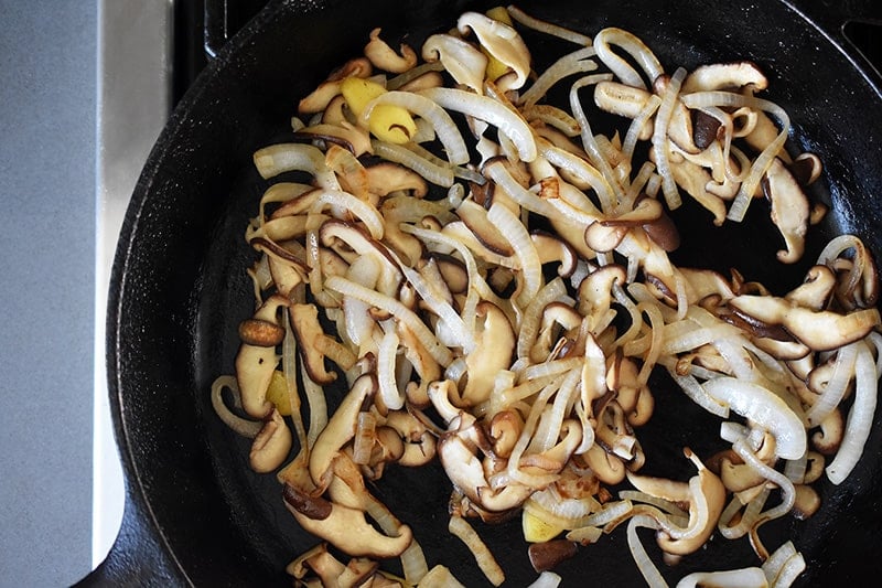 An overhead shot of onions, shiitake mushrooms, and ginger sautéing in a cast iron skillet.