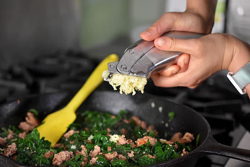 Minced garlic is added from a garlic press to the cooked sausage and kale in the cast iron skillet.