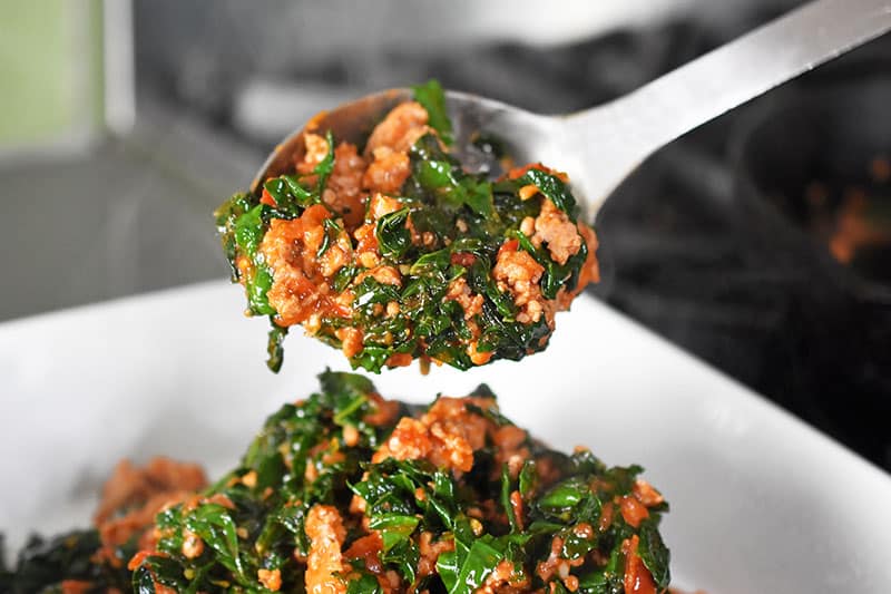 A large spoon transfers the cooked sausage, kale, and marinara to the greased casserole dish.