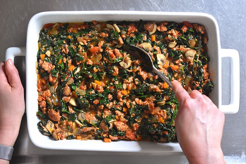 An overhead shot of someone mixing the Italian Sausage + Kale Casserole with a spoon.