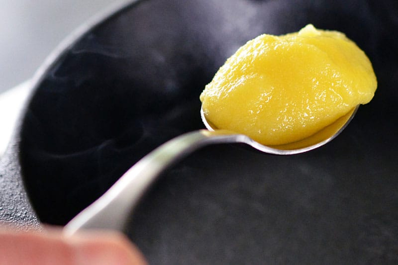 A spoon with ghee is hovering over a hot cast iron skillet.