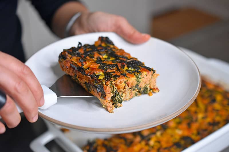 A square slice of Italian Sausage and Kale Casserole is being transferred to a white dish.