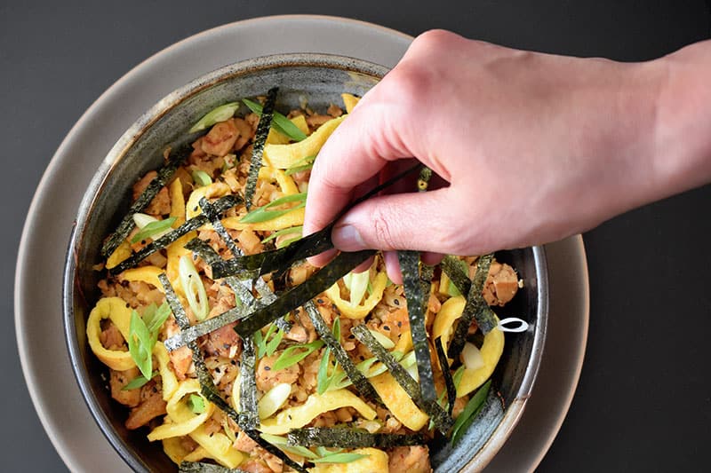 Someone adding slices of seaweed in a bowl with paleo kimchi cauliflower fried rice.
