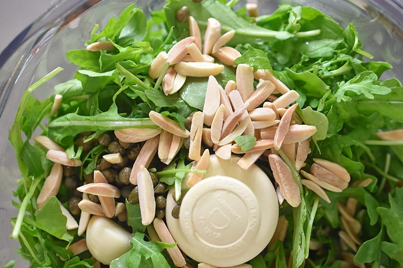 An overhead view of the ingredients for arugula pesto in a food processor.