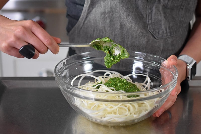 Someone adding a spoonful of paleo and Whole30 arugula pesto into a bowl of spiralized vegetables.