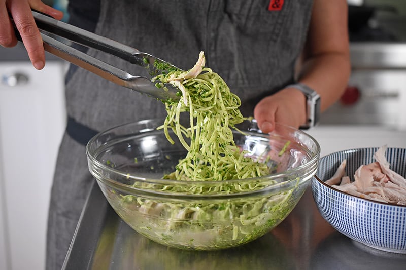 Someone mixing together zoodles, arugula pesto, and chicken in a bowl for a paleo and Whole30 dinner.